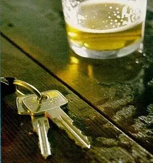 A set of car keys lies on a wet wooden surface next to a half-filled glass of beer.